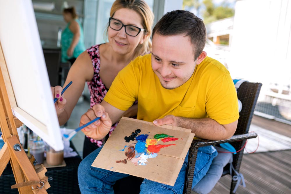 Ung glad kille med downs syndrom målar tillsammans med assistent